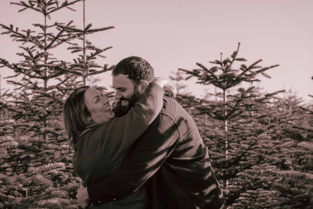 A couple embrace whilst playing in a Christmas tree plantation. The black and white image captures the love and frivolity of the moment.