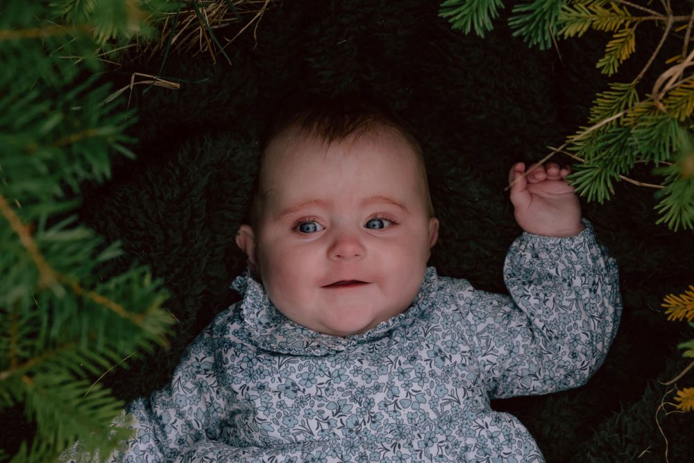 Adorable baby wearing a gray knit sweater smiles under green pine branches in soft lighting.