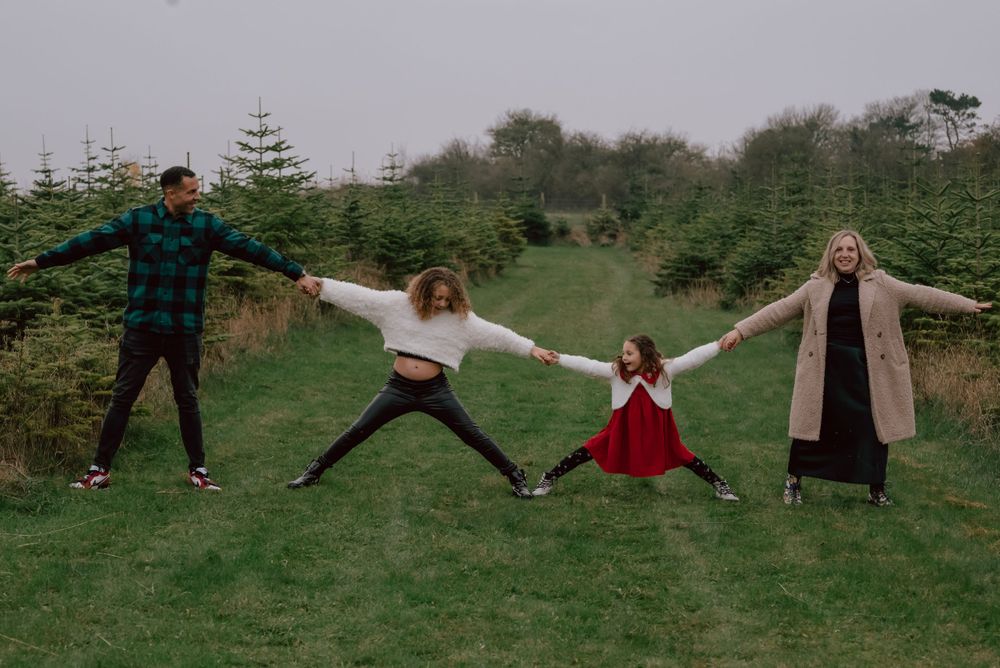 A group stands in a grassy field with outstretched arms in playful poses among evergreen trees on a cloudy day.