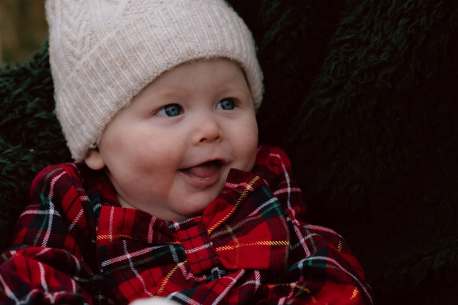 Smiling baby wearing a white knit beanie and red plaid winter coat.