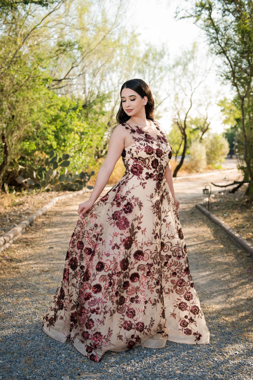 Woman in a floral burgundy ball gown poses elegantly on a sunlit outdoor path.