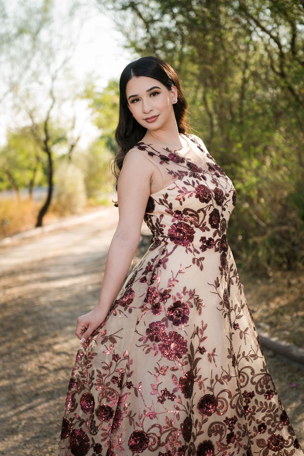 Woman in burgundy floral gown poses gracefully against natural outdoor backdrop.