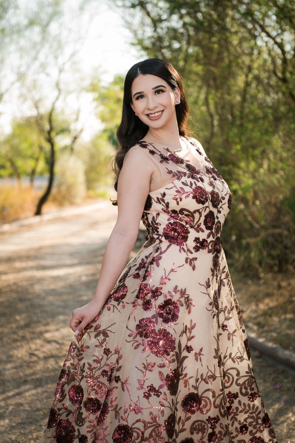 Woman wearing a cream and burgundy floral gown smiles while posing on a garden path.