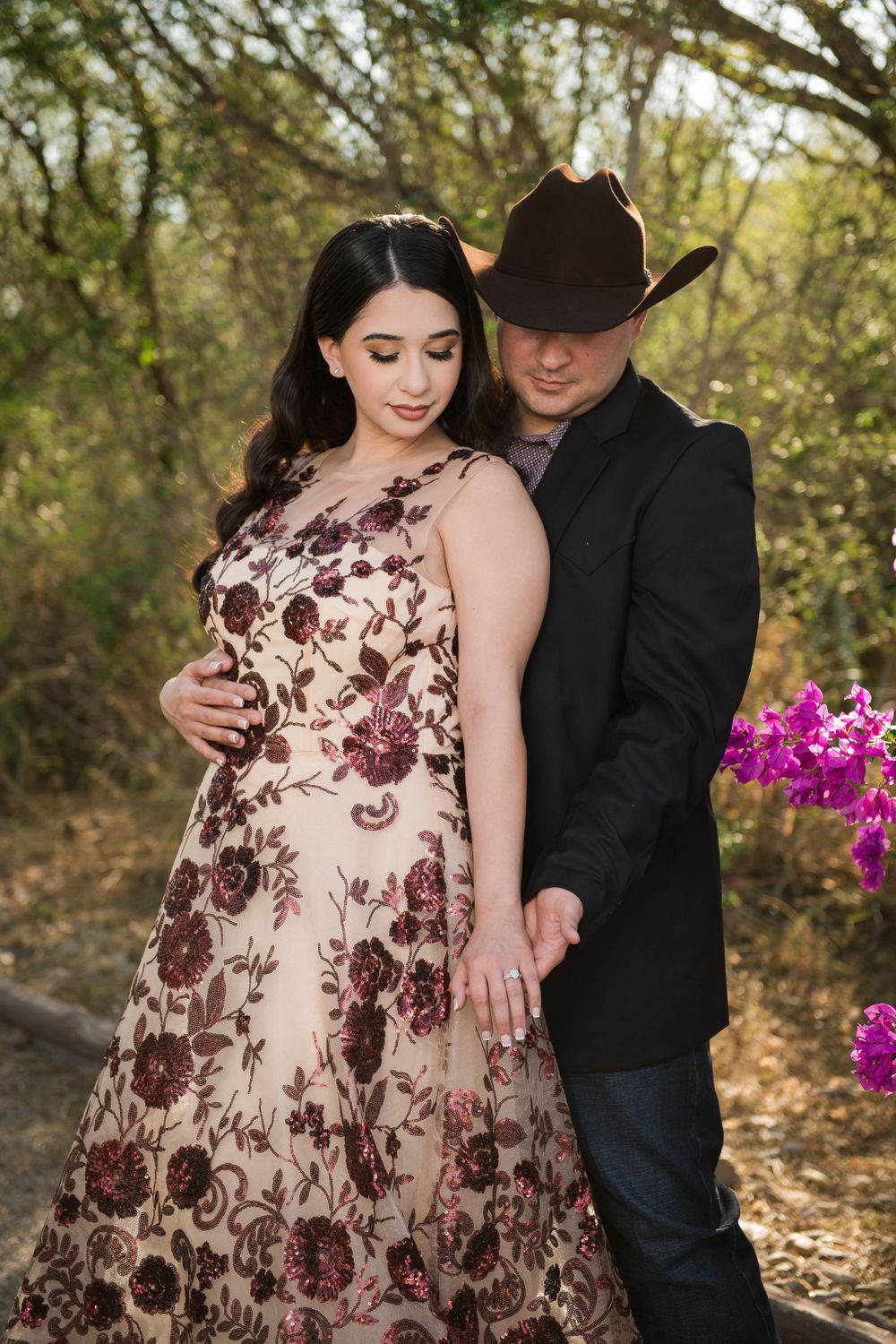 Couple poses intimately outdoors with her in floral dress and him in black with cowboy hat.