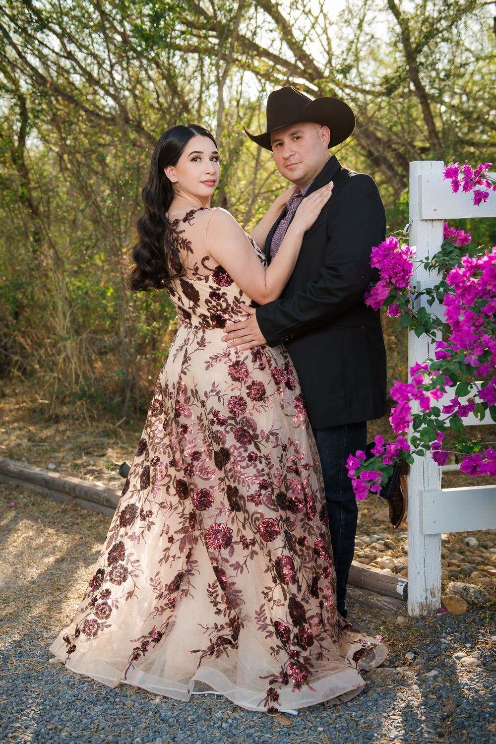Elegant couple embraces on garden path with purple flowers and white fence in background.