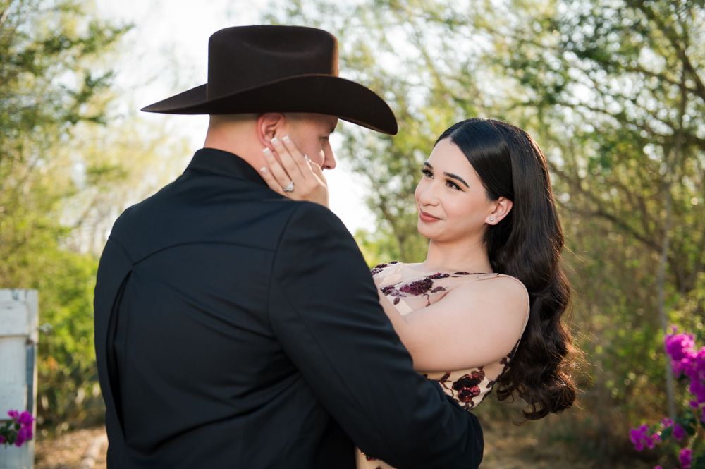 Couple embraces in front of desert foliage with the person in black wearing a cowboy hat and other in floral dress.