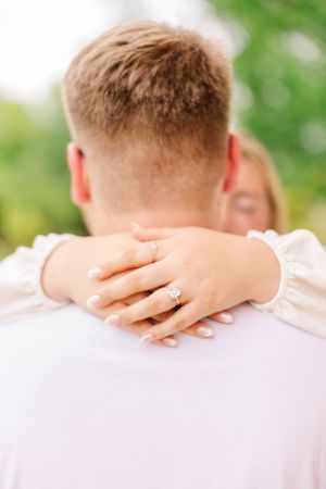 Close-up view of hands wearing engagement rings embracing around shoulders outdoors with green foliage background.