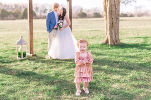A young child in a pink dress stands in front of a couple during their outdoor wedding.