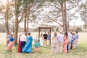 Guests gather for an intimate outdoor wedding ceremony under a wooden arch surrounded by pine trees.