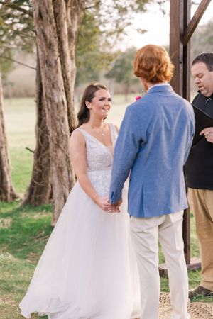 A couple exchanges vows during an outdoor wedding ceremony under towering trees.