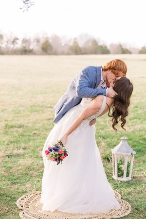 A romantic wedding couple shares a dip kiss in a field with a decorative lantern nearby.