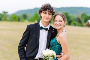 Couple in formal attire posing together at outdoor event with a bouquet and rural landscape in background.