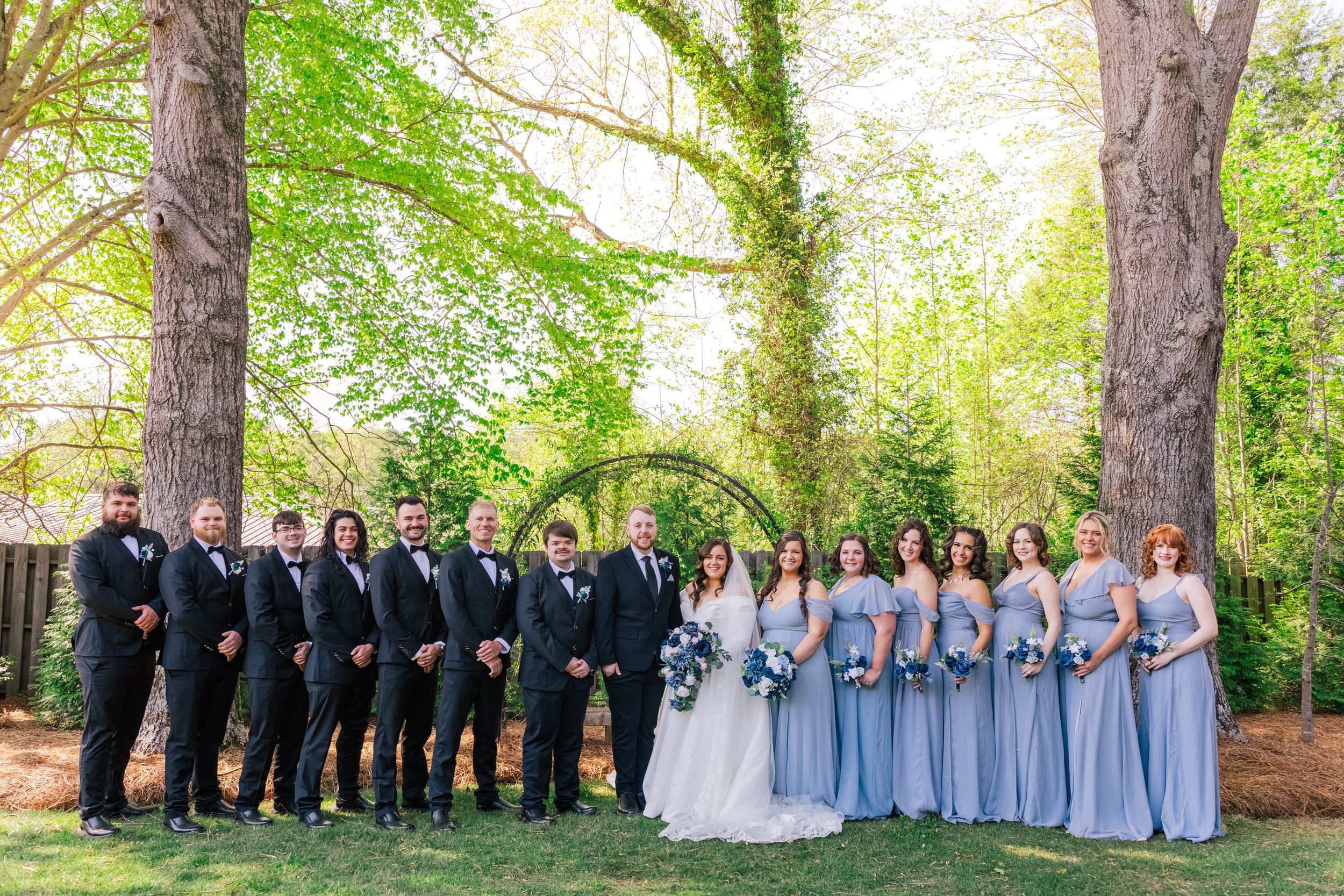 Large wedding party in blue dresses and dark suits posed in front of trees outdoors.
