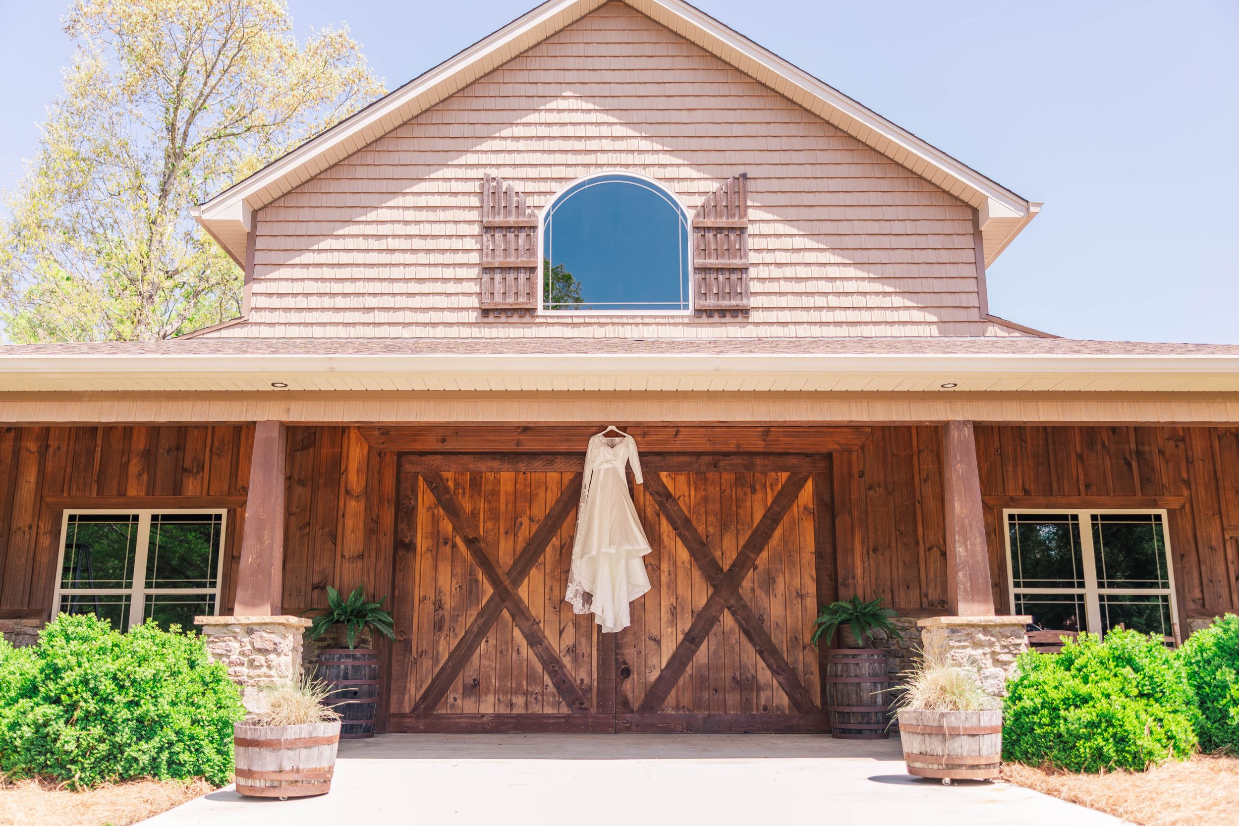 A white wedding dress hangs on rustic wooden barn doors of a blush-colored venue.