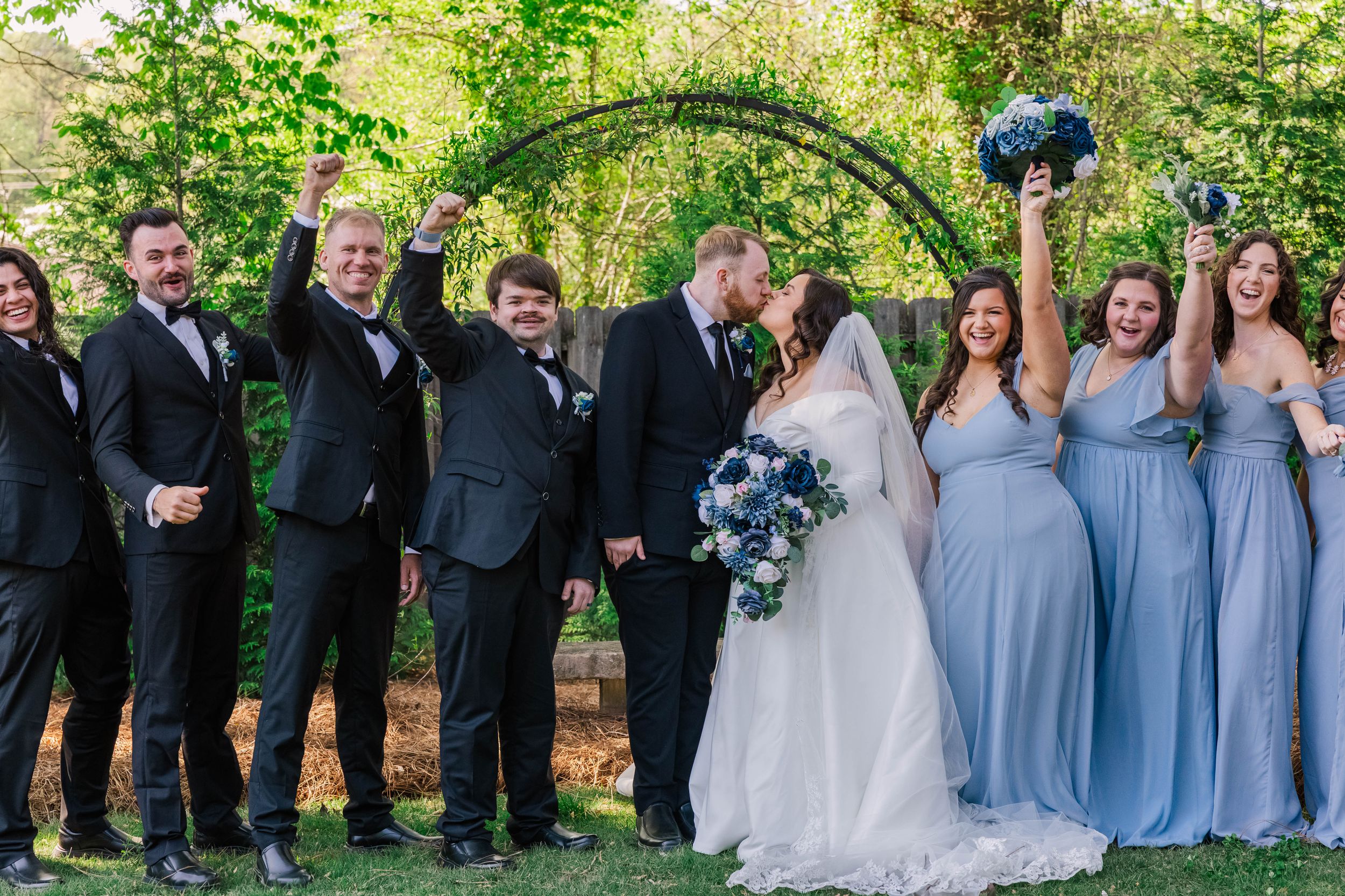 Wedding party members lined up under green trees wearing blue dresses and dark suits.