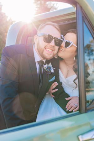 Newlyweds wearing sunglasses pose playfully in vintage car window with sun flare.