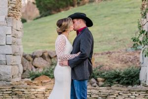 Couple shares intimate moment near rustic stone wall with bride wearing lace dress and groom in cowboy hat.