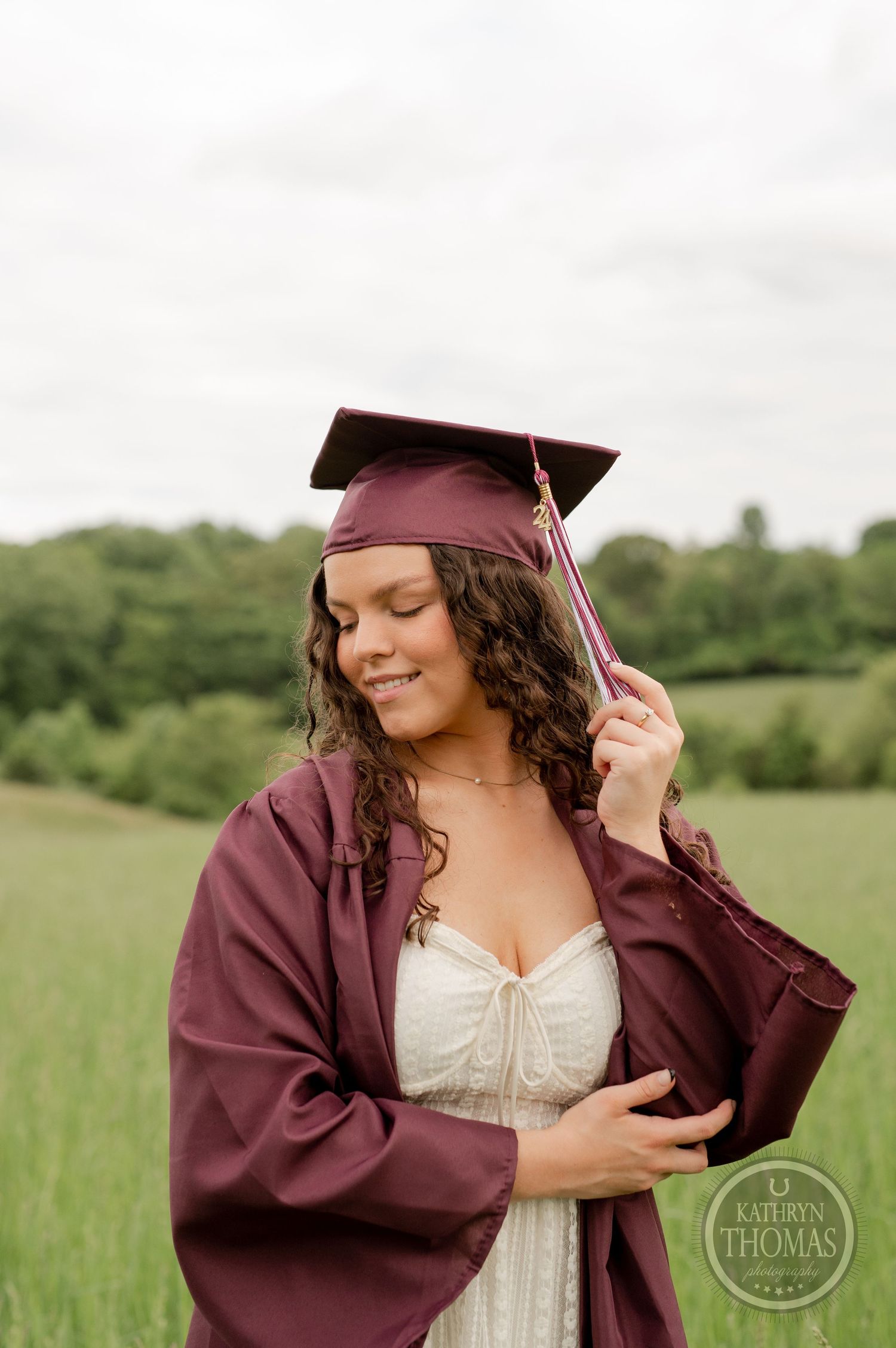 Graduate in maroon cap and gown with white dress poses thoughtfully in sunny field.