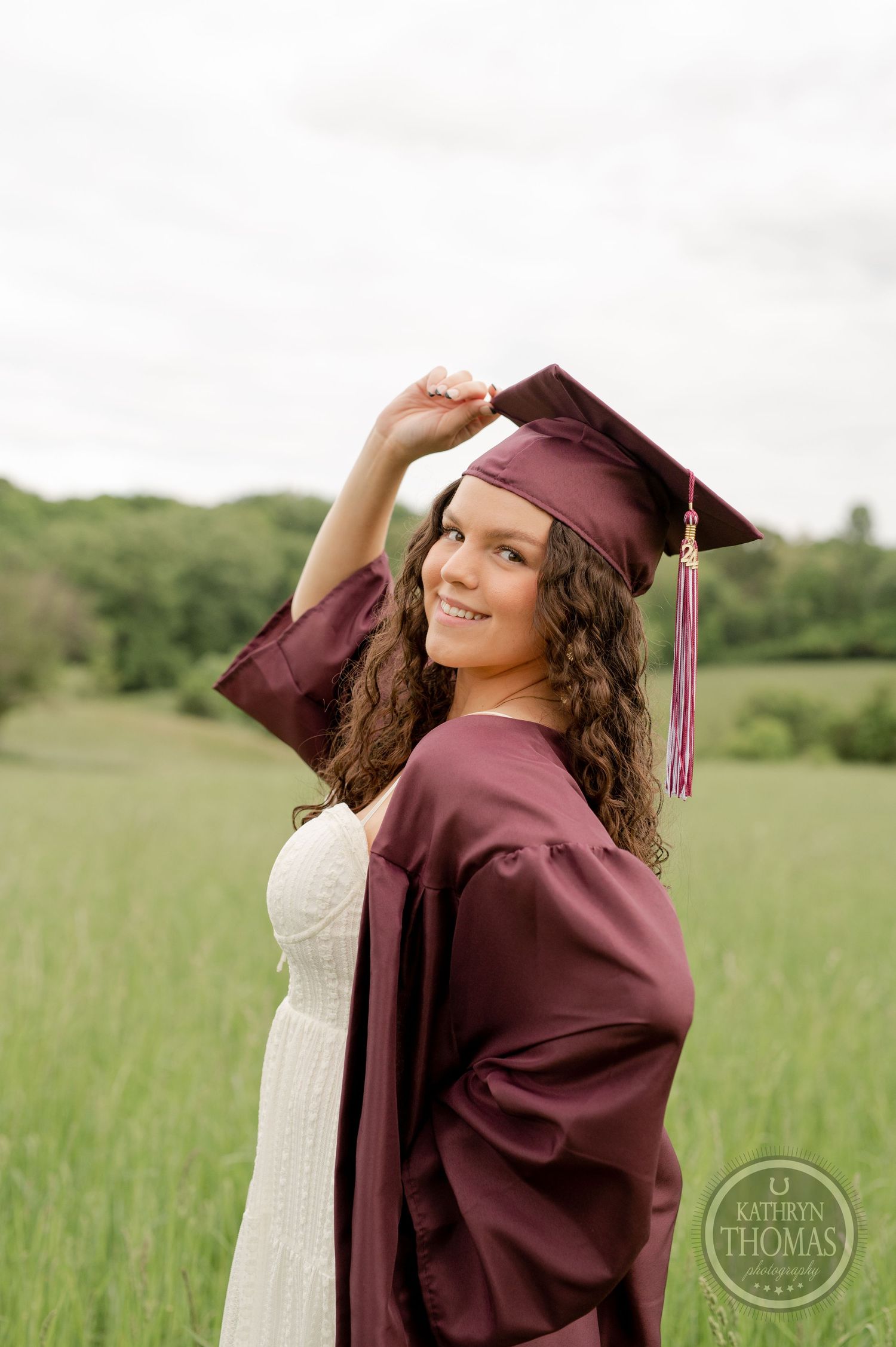 Graduate adjusts maroon cap while wearing white dress and graduation gown in outdoor field.