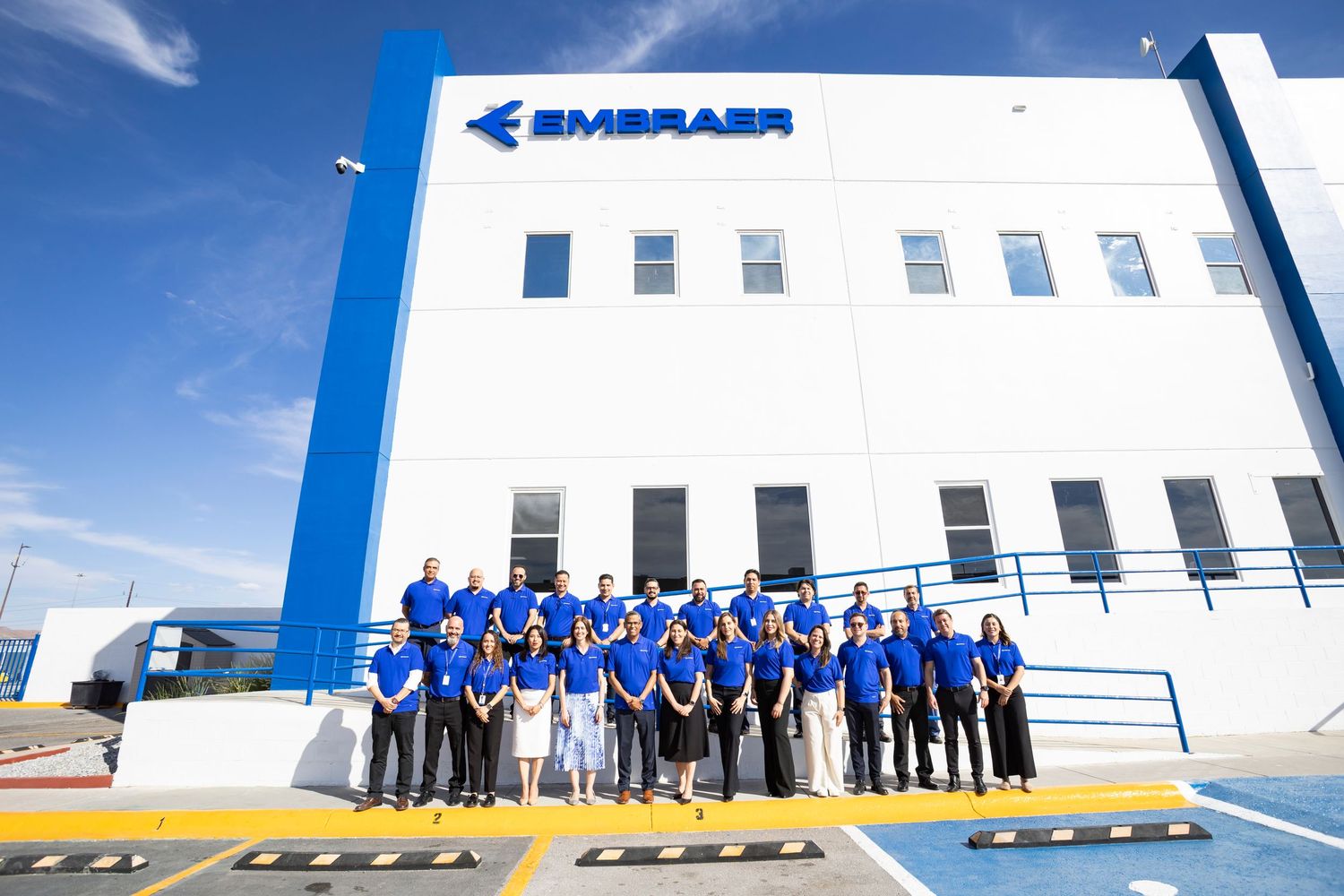 Group of Embraer employees in blue uniforms posing outside the Embraer headquarters building on a sunny day.