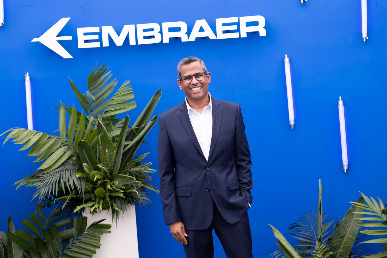 Man in suit smiling in front of Embraer blue logo wall with tropical plants.