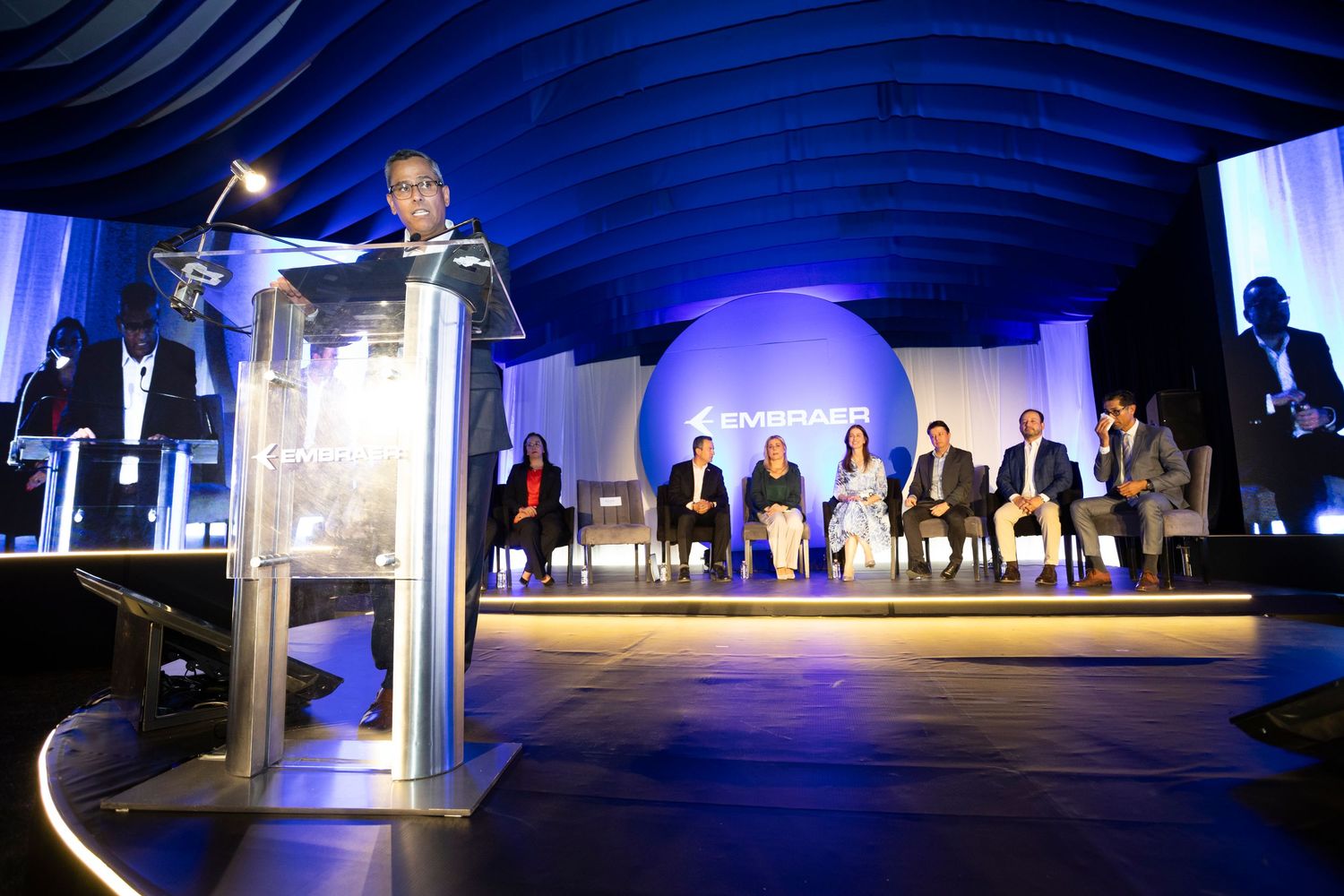 Speaker at podium addresses seated panel on blue-lit stage at formal corporate event or awards ceremony.