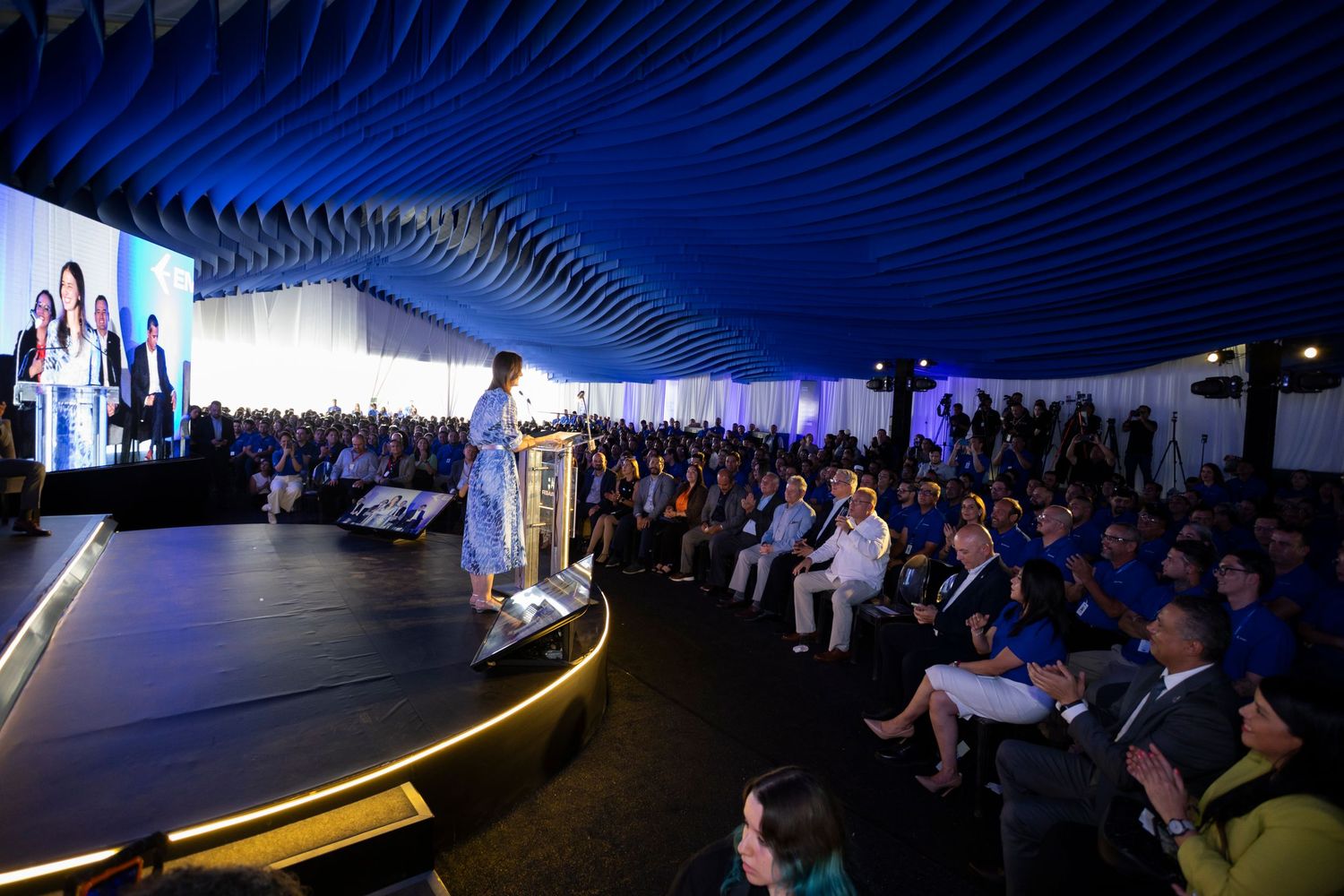 Speaker addresses large audience at formal blue-themed event under decorative ceiling with stage lighting.