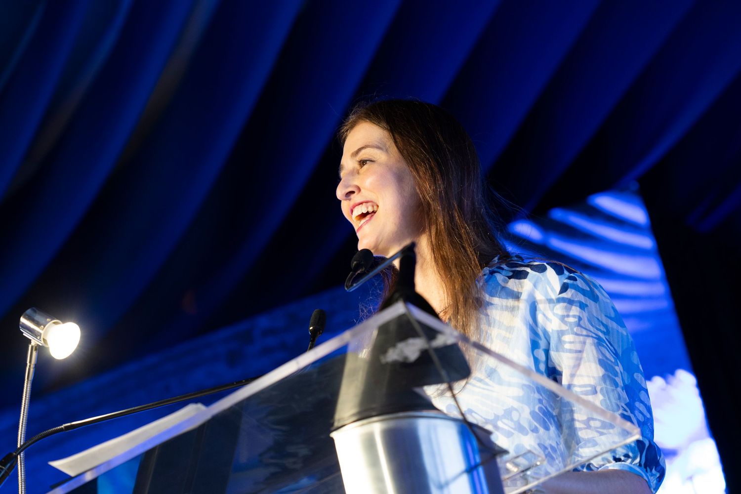 Young woman speaking at a glass podium under blue stage lighting at a formal event.