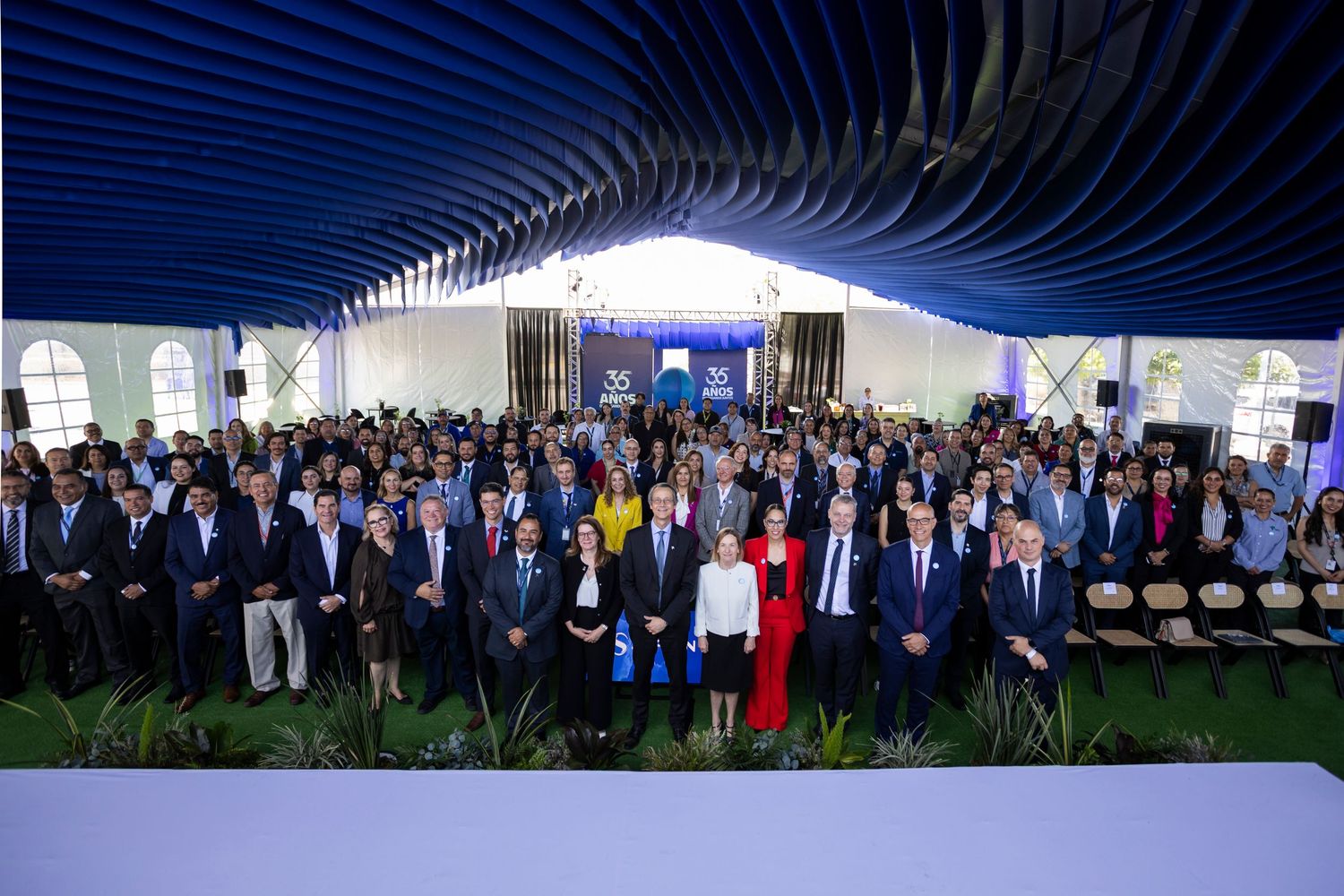 Large group of professionals posing together under a modern blue wave-patterned canopy at a formal corporate event.