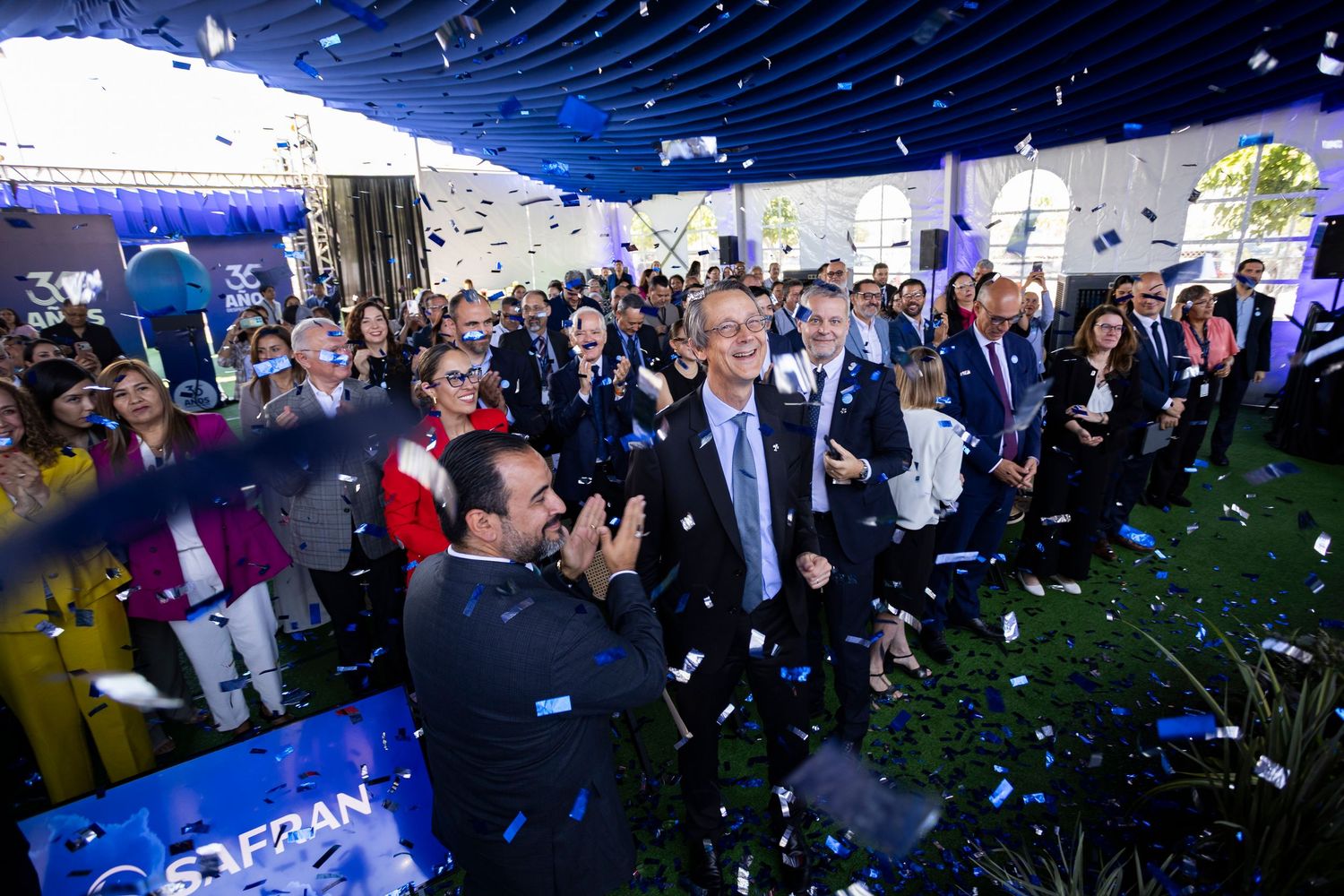 Crowd celebrating with blue and white confetti at a festive outdoor political event under a tent.