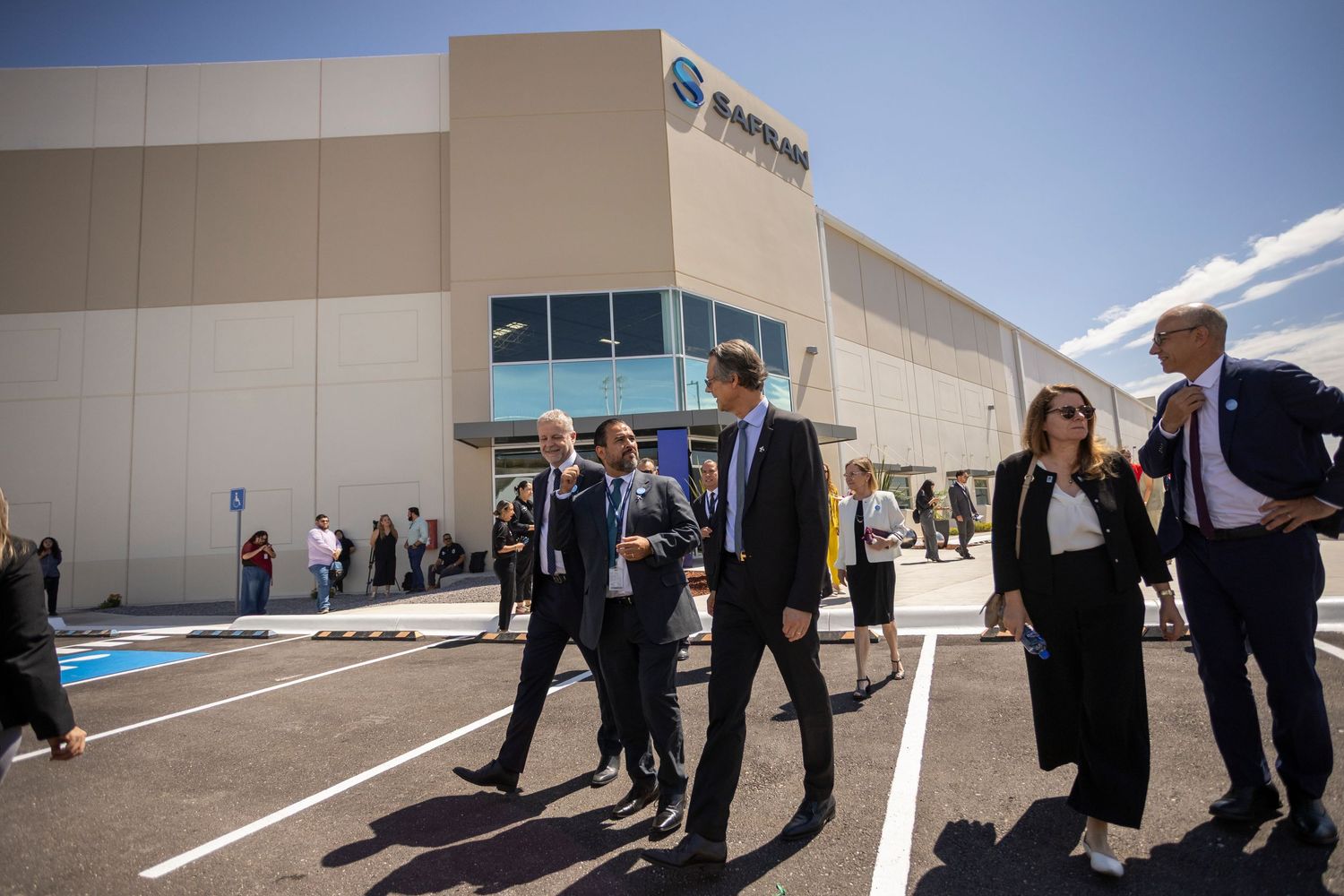 Business executives walk outside a modern industrial facility with a blue company logo on the building.