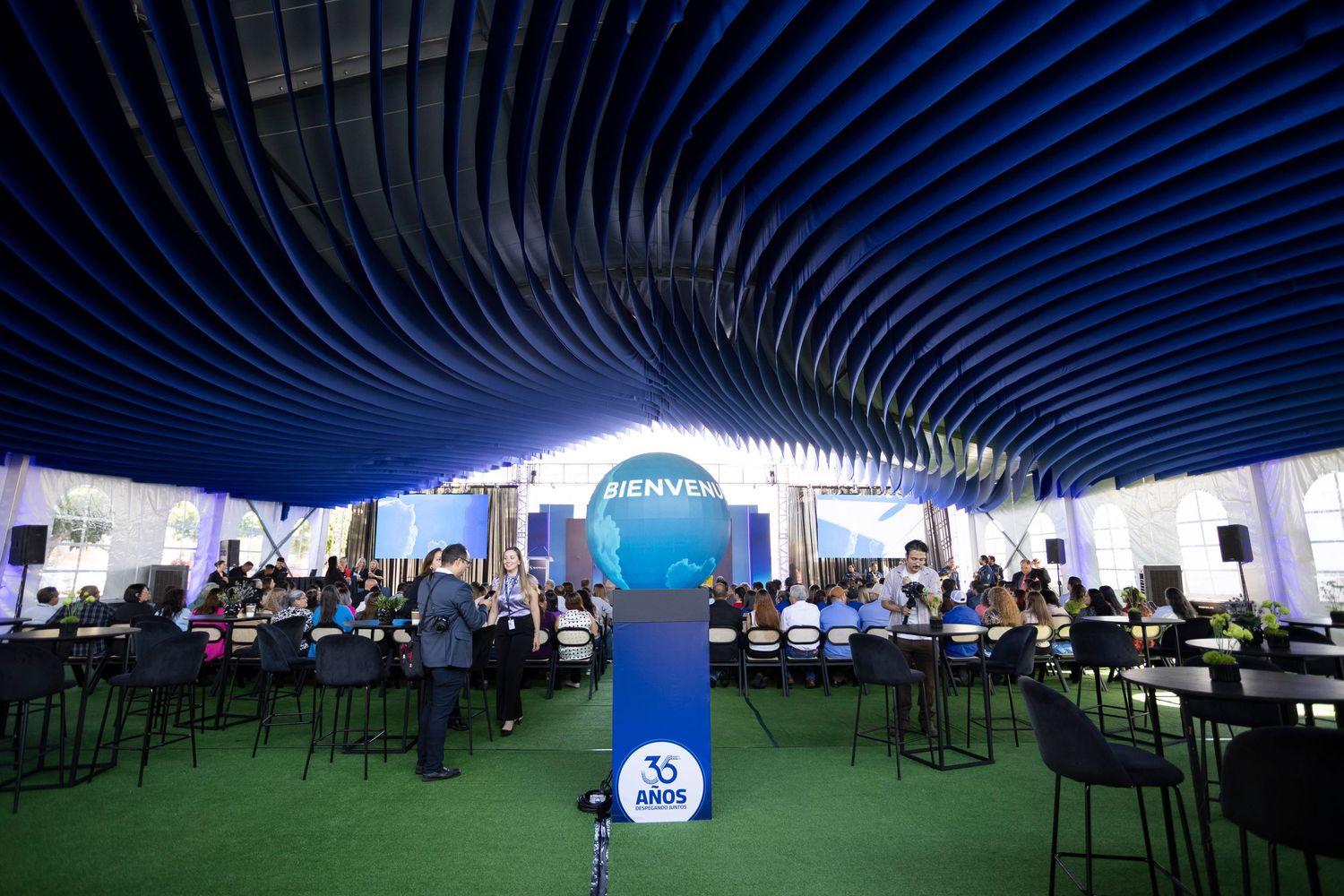 Corporate event venue with blue wave ceiling, globe centerpiece, and attendees seated at high tables under dramatic lighting.