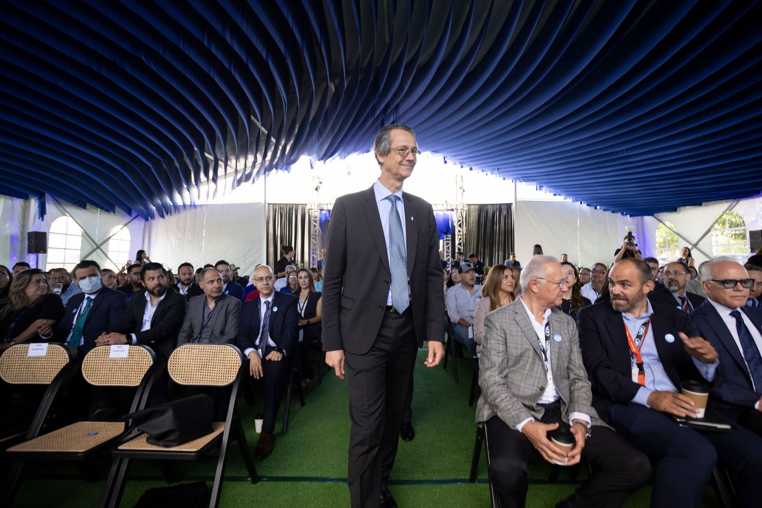 A tall man in a suit walks through rows of seated attendees under a blue tent at a formal outdoor event.