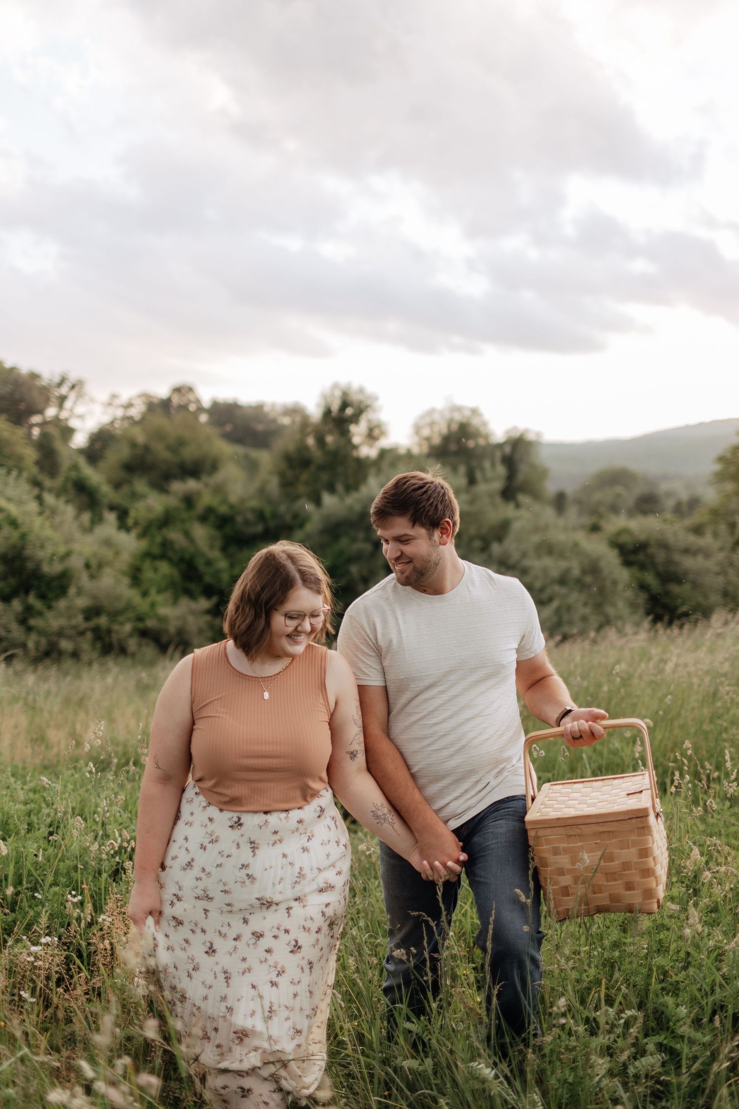 Casual couple walking through meadow with picnic basket at sunset.
