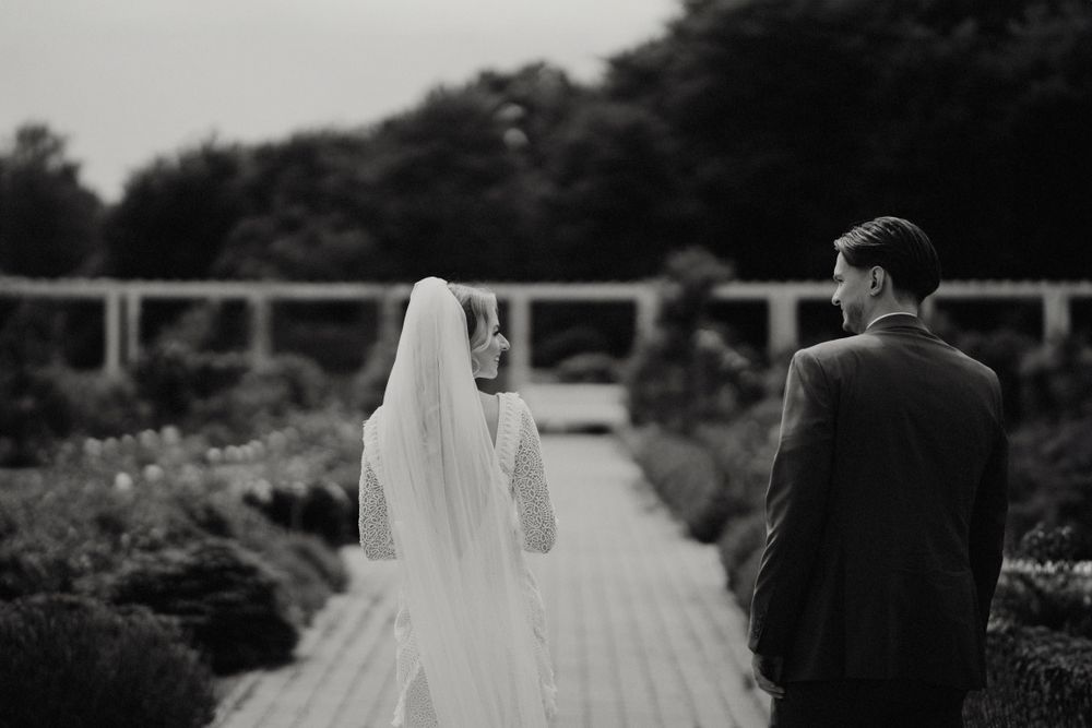 Newlyweds walk hand in hand through a garden path in black and white.