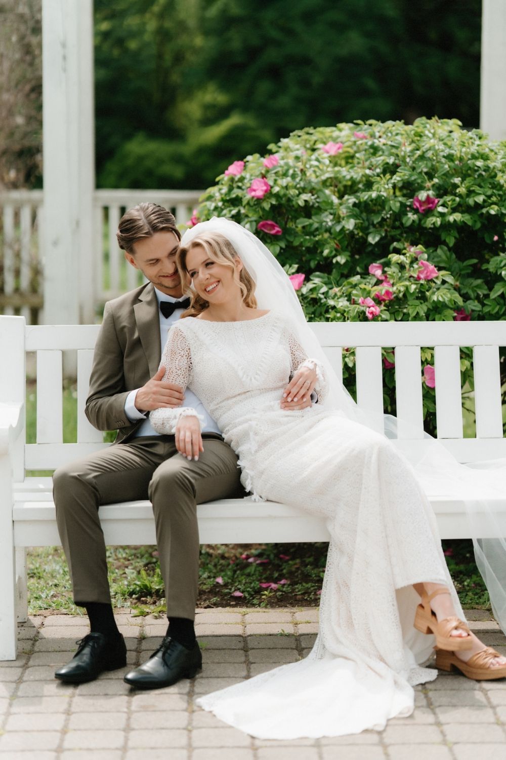 Couple shares tender moment on white garden bench surrounded by blooming roses.