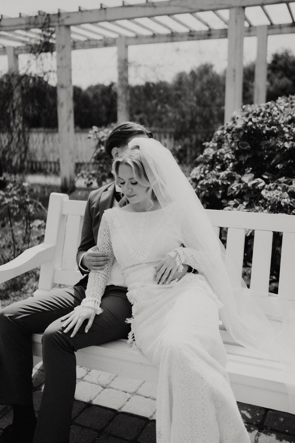 Black and white photo of newlyweds laughing together on a white pergola bench.