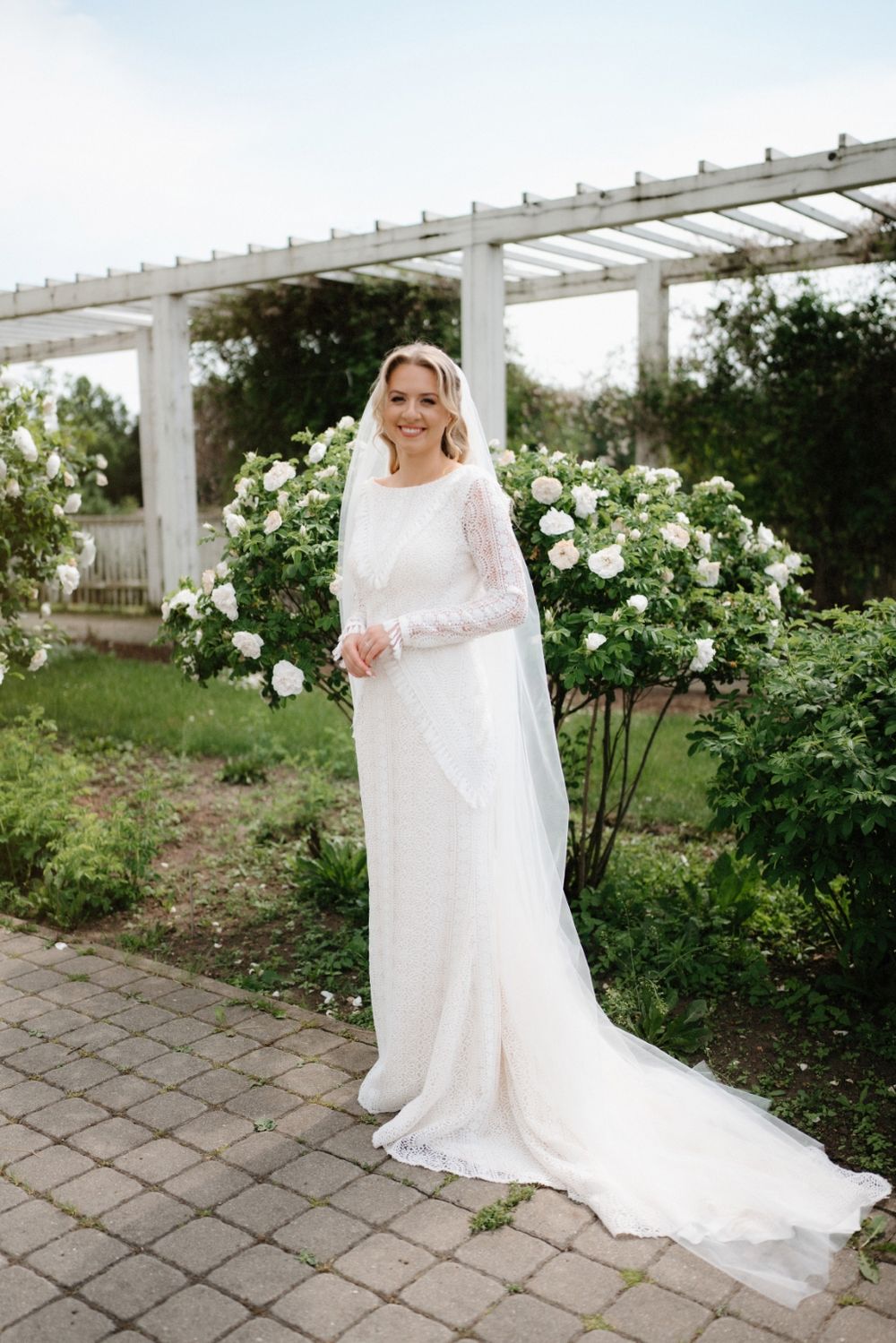 Bride in a flowing white dress standing near a white pergola in a garden with blooming roses.