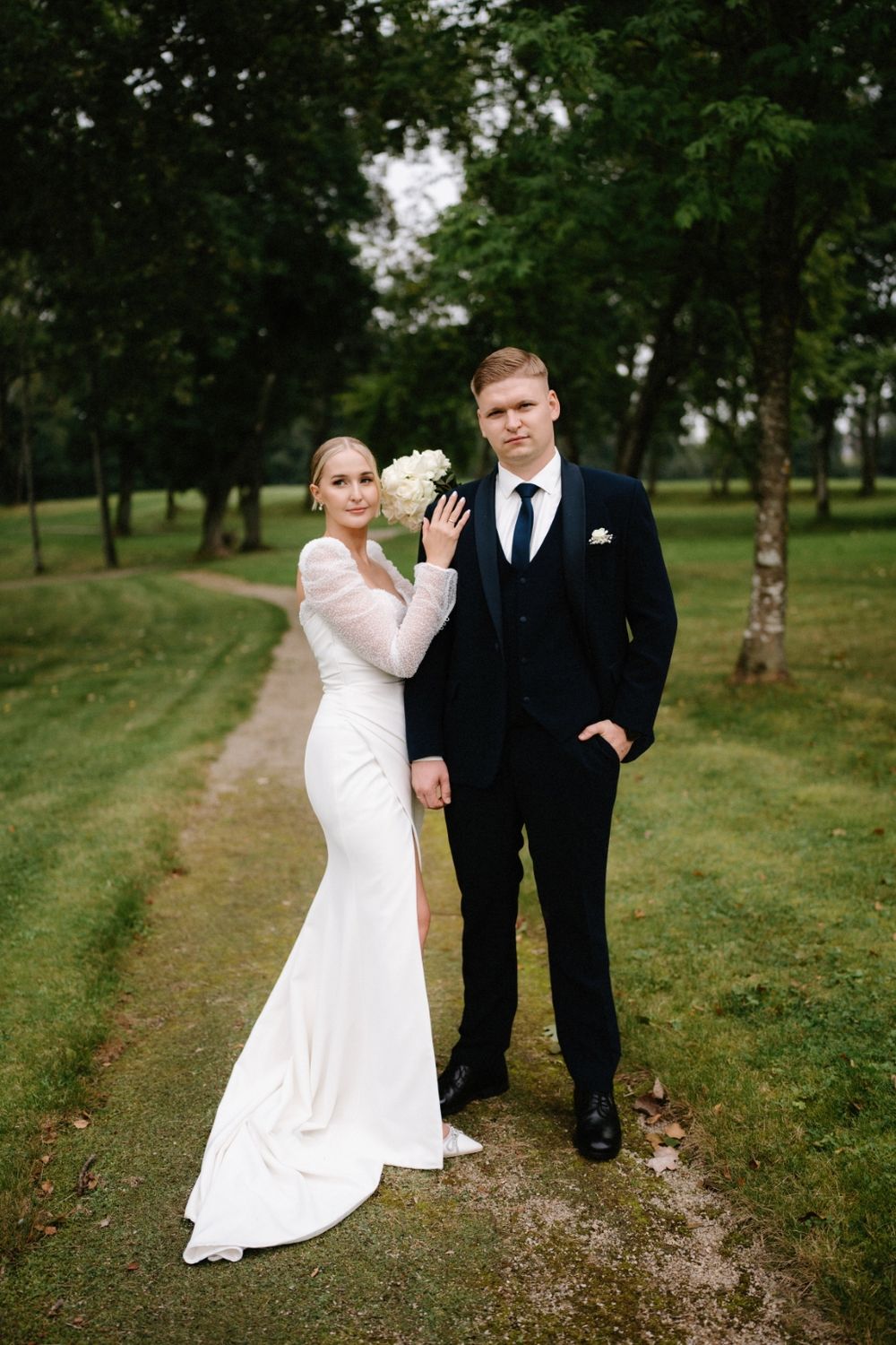 Bride and groom pose playfully on a gravel path surrounded by green trees and grass.