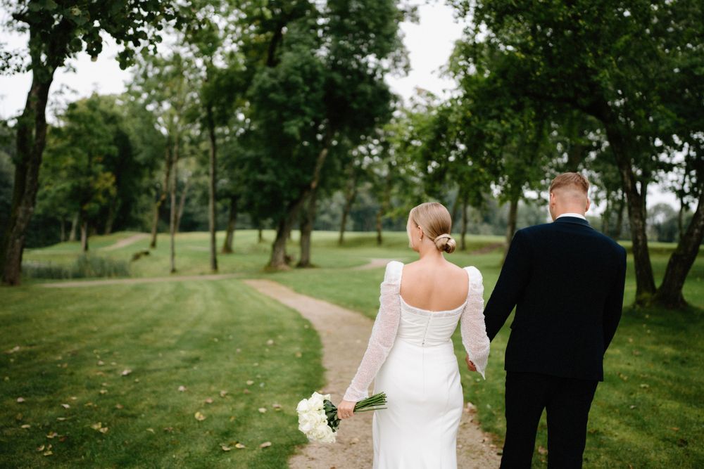 A couple walks hand in hand through a lush green park setting on their wedding day.
