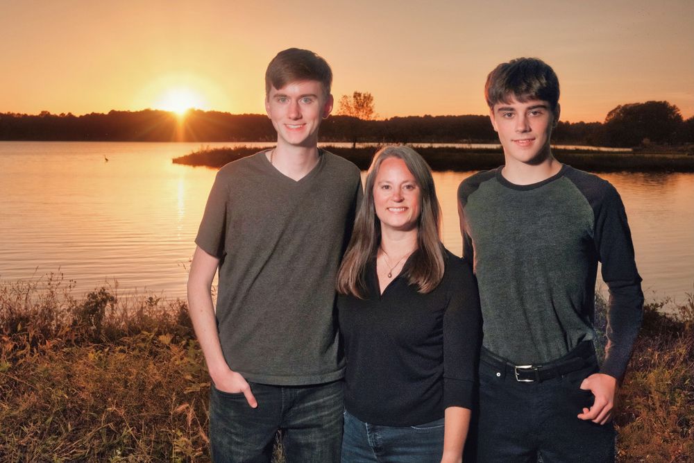Three people posing together at sunset by a lake with golden light creating silhouettes.