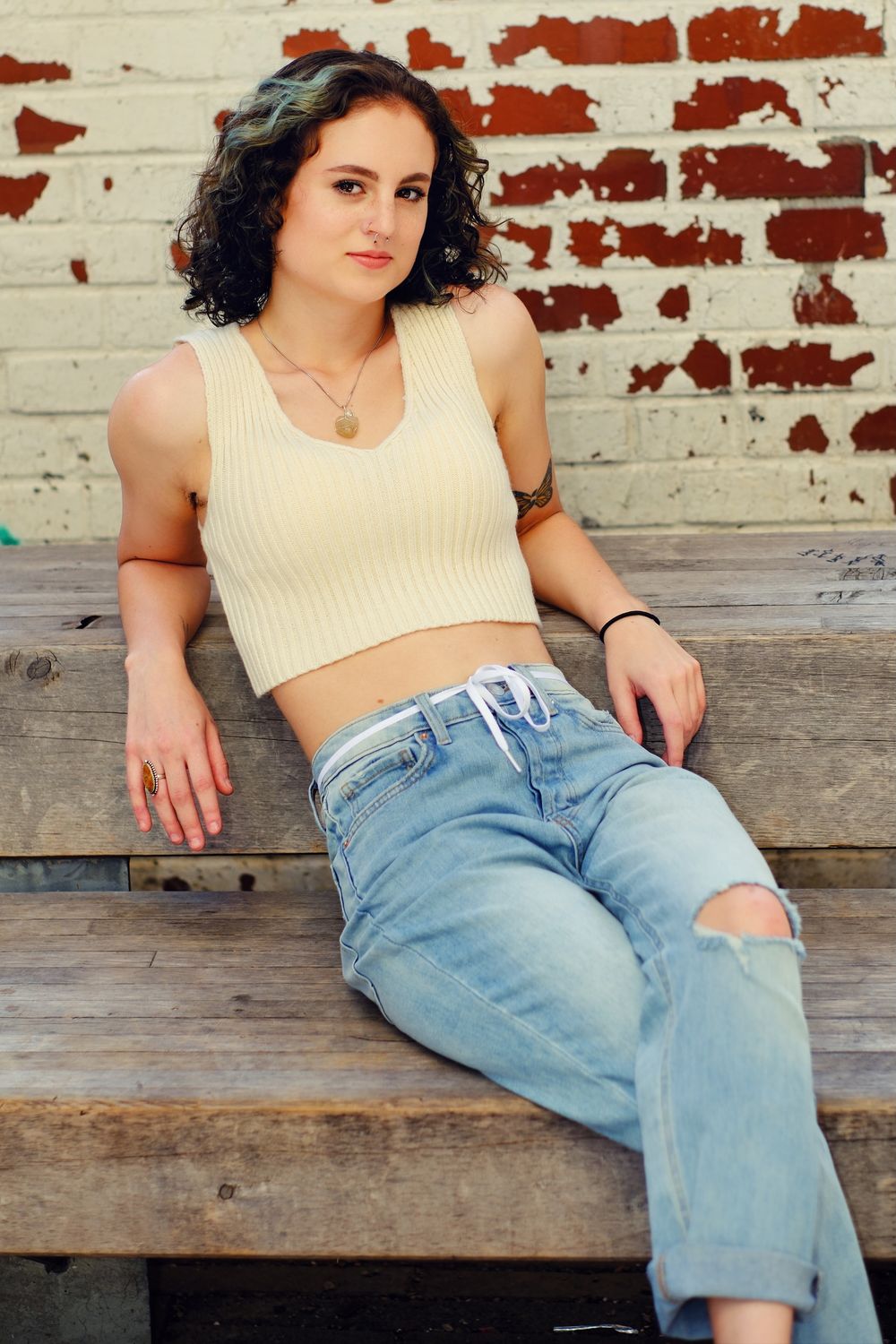 Portrait against weathered red brick wall wearing cream crop top and distressed jeans.