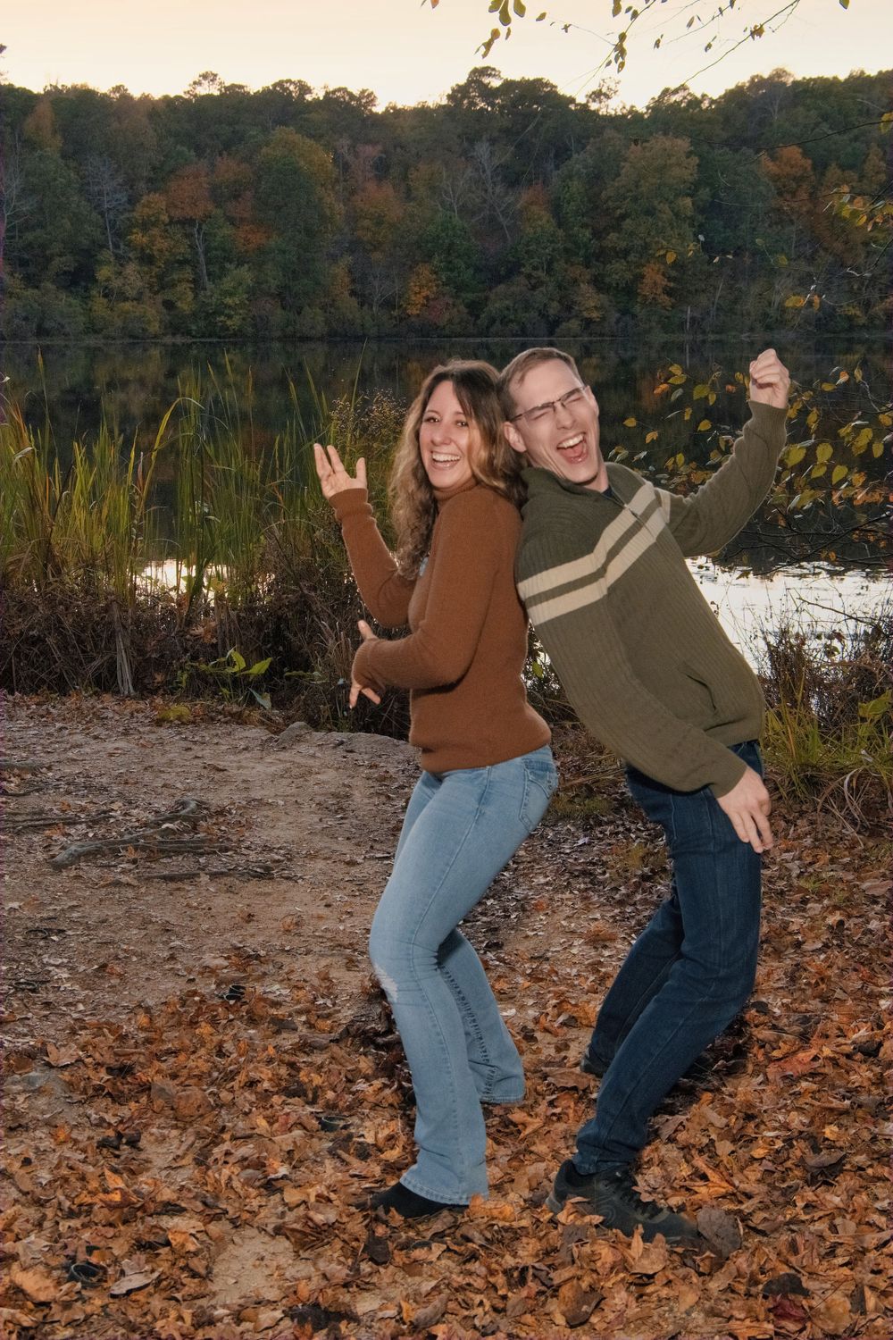 Two people posing playfully by a lake during autumn season.