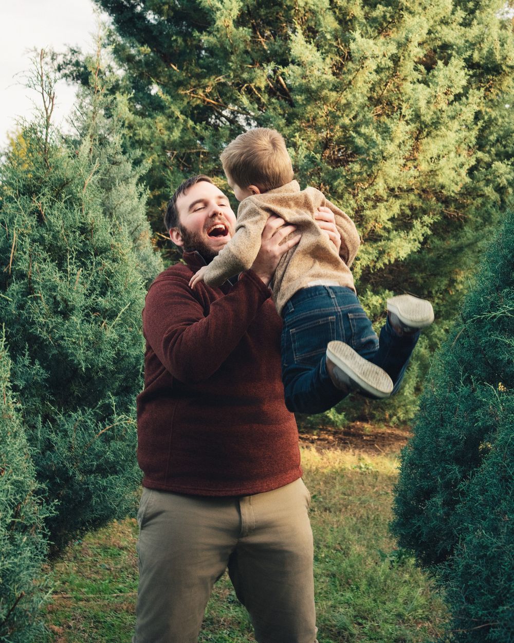 Playful moment of adult lifting child in air among evergreen trees.