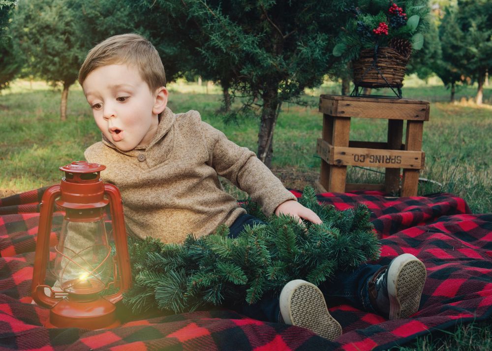 Child in brown sweater sits on plaid blanket with lantern in outdoor setting at twilight.