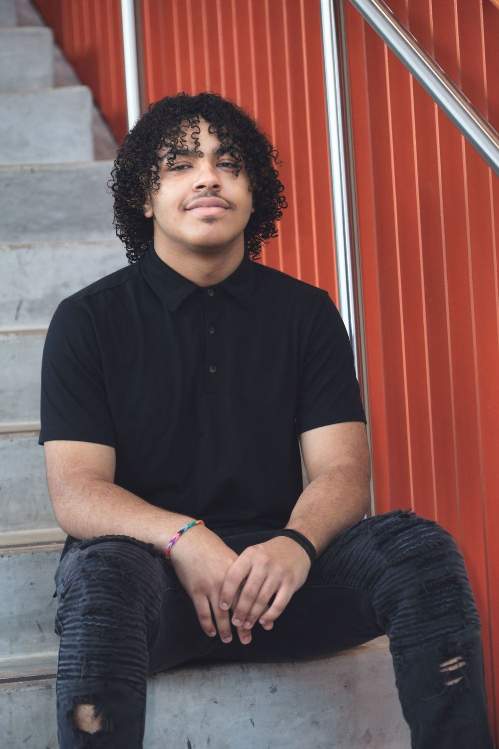Young person in black polo sits on concrete steps against red wall background.