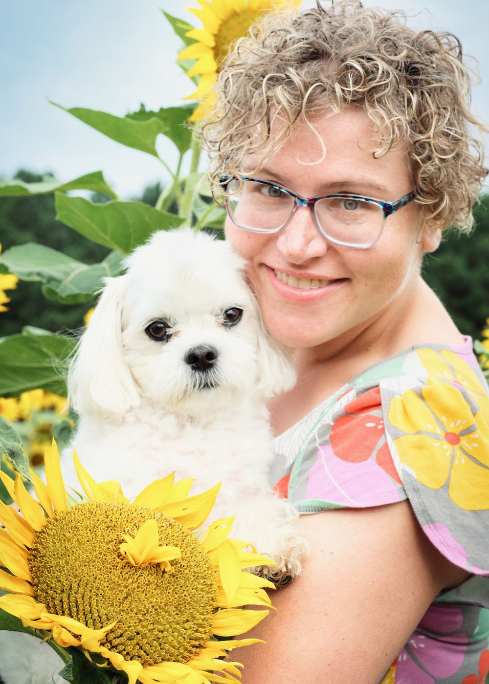 Person holds small white dog while posing with sunflowers in summer garden.