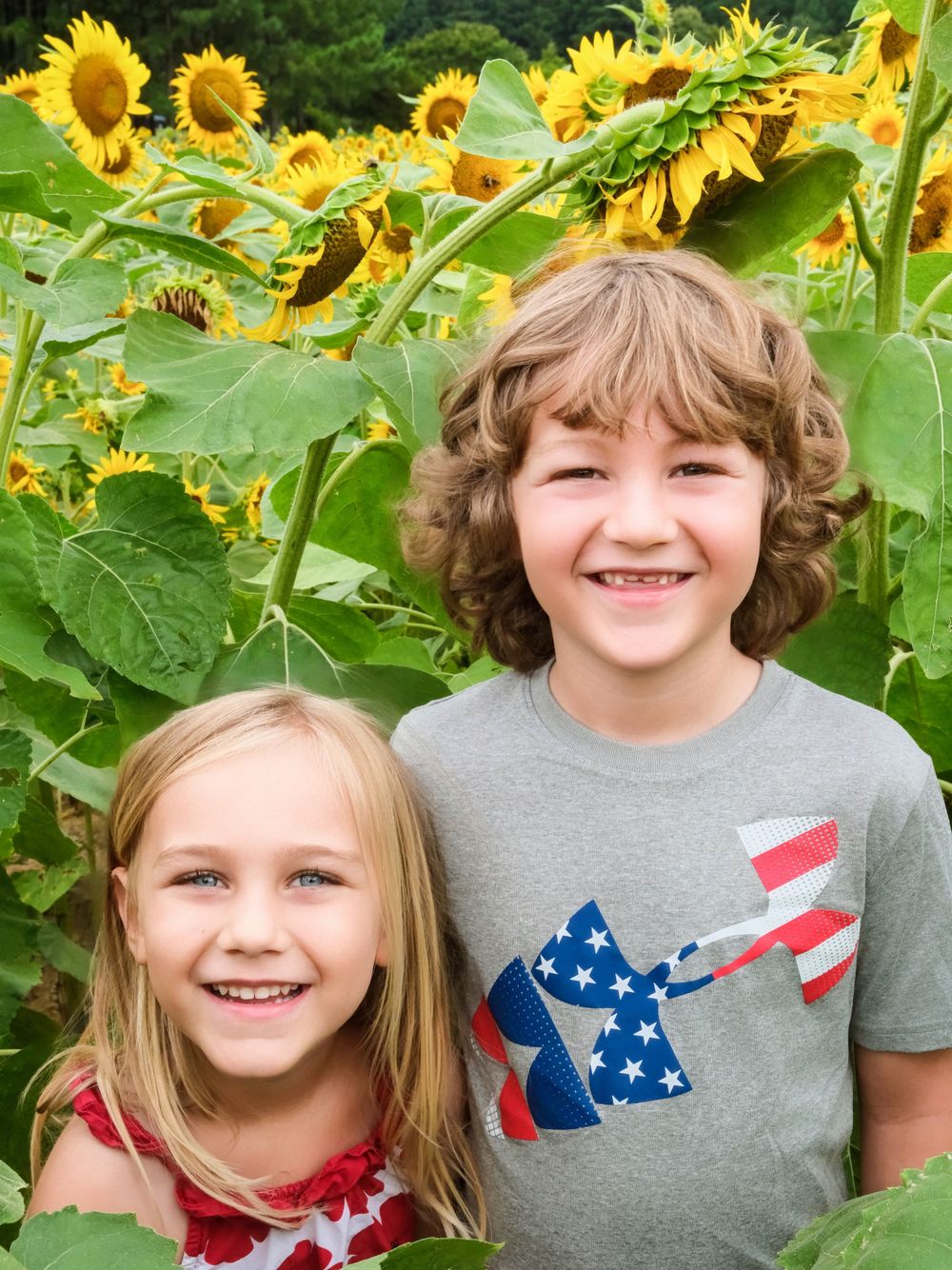 Two children smile together in front of tall blooming sunflower field.