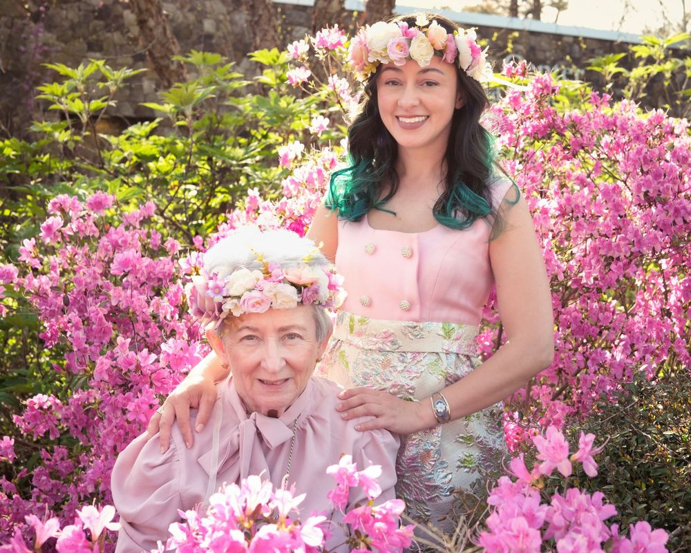 Two people wearing floral crowns pose together among blooming pink azalea bushes.