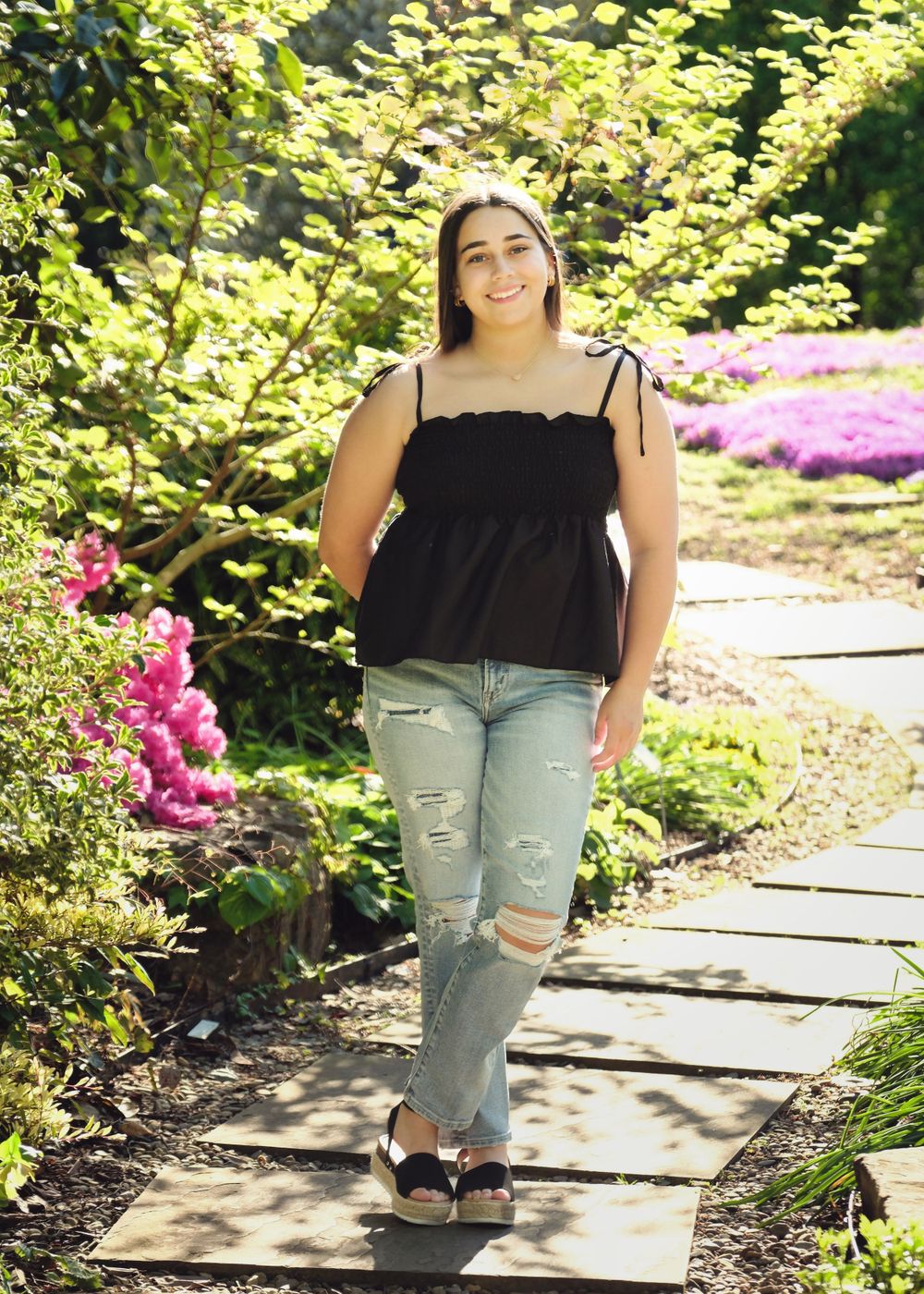 Person in black top poses on garden path surrounded by colorful flowers and greenery.
